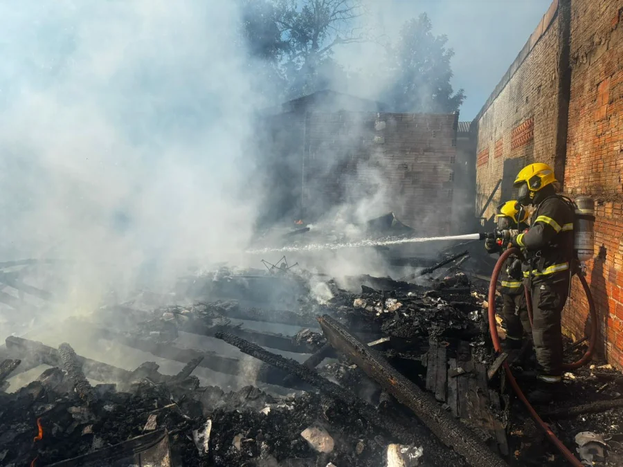 Dois bombeiros militares, utilizando equipamentos de proteção completos e capacetes amarelos, manuseiam uma mangueira para lançar água sobre os escombros fumegantes de uma residência completamente destruída pelo fogo. O chão está coberto por madeira carbonizada e destroços, com paredes de tijolos de edificações vizinhas visíveis ao fundo em meio à fumaça branca.