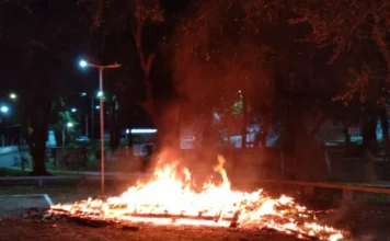 Incêndio destrói Casa do Papai Noel em Criciúma Fotografia noturna registra o momento em que a estrutura de madeira da "Casa do Papai Noel" é consumida por chamas intensas e laranjas. O fogo concentra-se na base da edificação de 8 m², situada em uma área aberta da Praça do Trabalhador. Ao fundo, é possível observar árvores e a iluminação pública do local sob o céu escuro da madrugada.