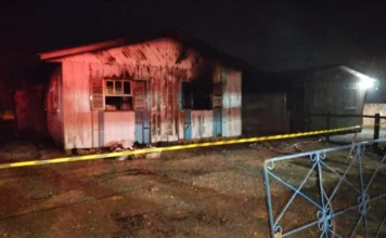 Incêndio destrói residência no bairro São Luis em Turvo Fotografia noturna mostra a fachada de uma residência de madeira de um pavimento, severamente danificada pelas chamas. As paredes brancas e azuis estão cobertas por fuligem e marcas de queimaduras profundas, especialmente ao redor das janelas. Em primeiro plano, uma fita de isolamento amarela da polícia atravessa a cena, e parte de uma grade metálica azul é visível à direita. O ambiente está escuro, iluminado apenas pelas luzes das viaturas de emergência.