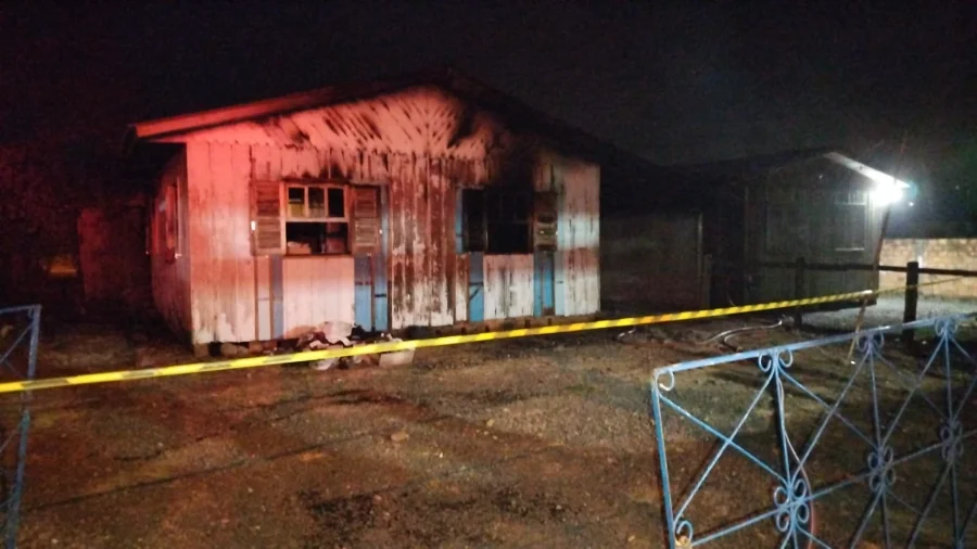 Fotografia noturna mostra a fachada de uma residência de madeira de um pavimento, severamente danificada pelas chamas. As paredes brancas e azuis estão cobertas por fuligem e marcas de queimaduras profundas, especialmente ao redor das janelas. Em primeiro plano, uma fita de isolamento amarela da polícia atravessa a cena, e parte de uma grade metálica azul é visível à direita. O ambiente está escuro, iluminado apenas pelas luzes das viaturas de emergência.