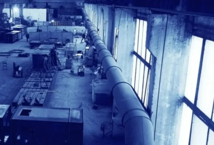 Indústria gaúcha teme perda de empregos com nova jornada Fotografia em perspectiva de um amplo pavilhão industrial com iluminação natural vinda de grandes janelas laterais. O cenário exibe maquinários pesados, tubulações metálicas extensas e trabalhadores ao fundo, representando o ambiente de produção da indústria de transformação no Rio Grande do Sul.