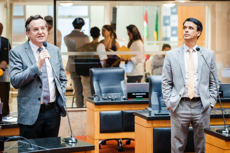 Fotografia em plano médio dos deputados Jair Miotto e Marcos da Rosa no plenário da Assembleia Legislativa de Santa Catarina (Alesc). À esquerda, o deputado Jair Miotto, de óculos e terno cinza escuro, fala ao microfone. À direita, o deputado Marcos da Rosa, vestindo terno cinza claro e gravata dourada, acompanha o pronunciamento. Ao fundo, é possível ver a estrutura do plenário com divisórias de vidro e outras pessoas em atividade.