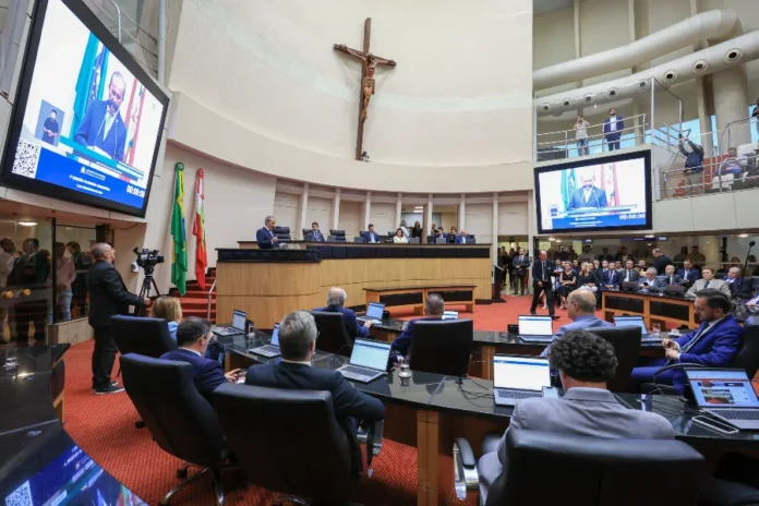 Jorginho Mello abre ano legislativo de 2026 na Alesc Fotografia do Plenário Deputado Osni Régis, na Assembleia Legislativa de Santa Catarina (Alesc), durante a sessão especial de abertura dos trabalhos legislativos de 2026. O governador Jorginho Mello aparece discursando no púlpito central e sua imagem é transmitida em dois grandes telões localizados nas laterais do plenário. Deputados e outras autoridades estão sentados nas bancadas, acompanhando a leitura da Mensagem Anual.