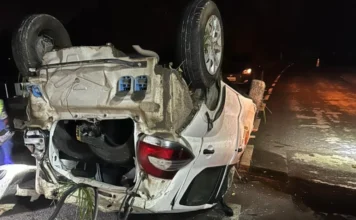 Jovem fica ferido após capotamento na SC-108 em Urussanga Fotografia noturna em plano médio de um Fiat Mobi branco capotado sobre o asfalto da rodovia SC-108. O veículo está com as quatro rodas voltadas para cima, apresentando danos na lataria e restos de vegetação presos à estrutura inferior. Ao fundo, luzes de sinalização e de outro veículo iluminam a pista escura, evidenciando o cenário do acidente no bairro Rio Maior.