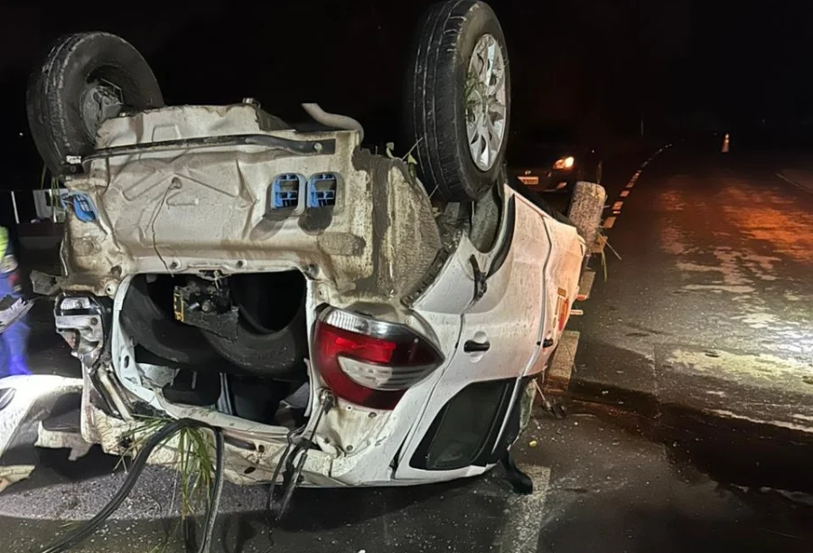 Jovem fica ferido após capotamento na SC-108 em Urussanga Fotografia noturna em plano médio de um Fiat Mobi branco capotado sobre o asfalto da rodovia SC-108. O veículo está com as quatro rodas voltadas para cima, apresentando danos na lataria e restos de vegetação presos à estrutura inferior. Ao fundo, luzes de sinalização e de outro veículo iluminam a pista escura, evidenciando o cenário do acidente no bairro Rio Maior.