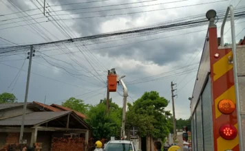 Jovem morre eletrocutado ao tentar desligar disjuntor Registro da operação de socorro e isolamento de área no bairro Cristo Redentor, em Criciúma. A imagem mostra bombeiros fardados e populares em uma rua residencial sob céu nublado. Ao centro, um técnico em um cesto elevatório de um caminhão branco realiza reparos na fiação elétrica de um poste, enquanto uma viatura do Corpo de Bombeiros (CBMSC) é visível em primeiro plano, à direita.