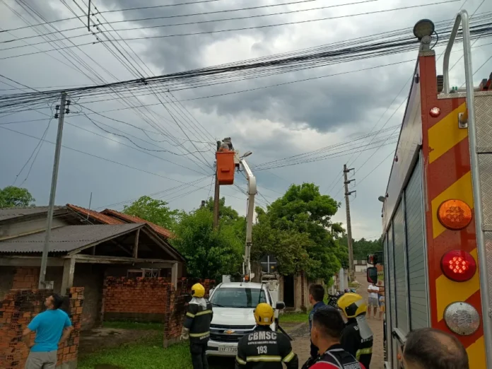 Jovem morre eletrocutado ao tentar desligar disjuntor Registro da operação de socorro e isolamento de área no bairro Cristo Redentor, em Criciúma. A imagem mostra bombeiros fardados e populares em uma rua residencial sob céu nublado. Ao centro, um técnico em um cesto elevatório de um caminhão branco realiza reparos na fiação elétrica de um poste, enquanto uma viatura do Corpo de Bombeiros (CBMSC) é visível em primeiro plano, à direita.