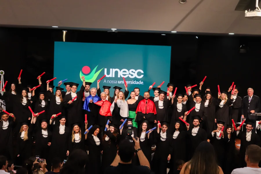Grupo de formandos da Unesc, incluindo a turma de Direito, celebra no palco levantando os canudos durante a cerimônia de colação de grau em Criciúma, onde a estudante Larissa Vidal Borba recebeu seu diploma após superar um câncer raro.