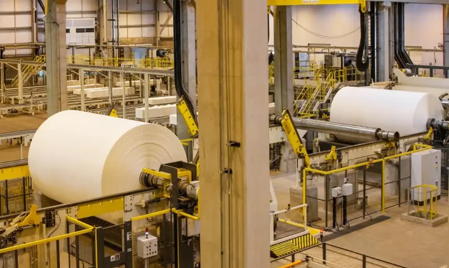 Vista interna de uma fábrica de papel com grandes rolos em produção e maquinário industrial. A imagem ilustra o setor produtivo brasileiro, que segundo a Confederação Nacional da Indústria (CNI) e dados do IBGE, enfrentou estagnação em 2025 devido aos altos juros da Taxa Selic e ao aumento das importações, resultando em um crescimento modesto de apenas 0,6% no ano.