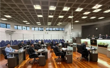 Legislativo de Araranguá inicia atividades de 2026 Fotografia do Plenário Altícimo Tournier durante a primeira sessão ordinária de 2026 da Câmara de Araranguá. A imagem mostra os vereadores sentados em suas bancadas de madeira, organizadas em semicírculo sobre um piso de madeira polida. Ao fundo, vê-se a Mesa Diretora composta por quatro membros e as galerias destinadas ao público, protegidas por divisórias de vidro. O ambiente é amplo, com iluminação embutida no teto quadriculado.