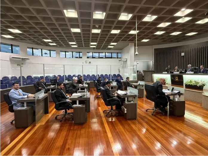 Legislativo de Araranguá inicia atividades de 2026 Fotografia do Plenário Altícimo Tournier durante a primeira sessão ordinária de 2026 da Câmara de Araranguá. A imagem mostra os vereadores sentados em suas bancadas de madeira, organizadas em semicírculo sobre um piso de madeira polida. Ao fundo, vê-se a Mesa Diretora composta por quatro membros e as galerias destinadas ao público, protegidas por divisórias de vidro. O ambiente é amplo, com iluminação embutida no teto quadriculado.