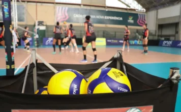 Mampituba/Unesc enfrenta Asa Alumínio pela Superliga B em SP Carrinho contendo bolas de vôlei em primeiro plano, com atletas da equipe feminina do Mampituba treinando em quadra ao fundo, desfocadas, durante preparação para partida da Superliga B.