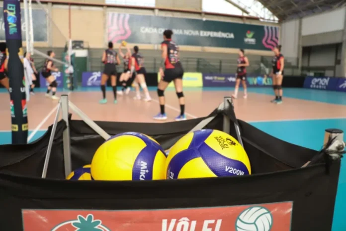 Mampituba Unesc enfrenta Asa Alumínio pela Superliga B em SP Carrinho contendo bolas de vôlei em primeiro plano, com atletas da equipe feminina do Mampituba treinando em quadra ao fundo, desfocadas, durante preparação para partida da Superliga B.