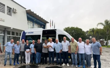 Meleiro recebe nova van para transporte de pacientes Grupo de autoridades municipais de Meleiro posa para foto em frente a uma van branca de transporte de saúde. O veículo está estacionado em um pátio de pedras portuguesas, diante do prédio da Prefeitura. No centro da imagem, dois homens seguram as chaves do automóvel em um ato simbólico de entrega. A van possui adesivos de identificação da Secretaria de Saúde e do serviço de transporte sanitário.