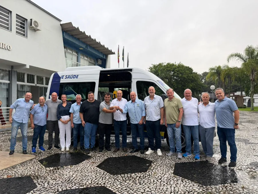 Grupo de autoridades municipais de Meleiro posa para foto em frente a uma van branca de transporte de saúde. O veículo está estacionado em um pátio de pedras portuguesas, diante do prédio da Prefeitura. No centro da imagem, dois homens seguram as chaves do automóvel em um ato simbólico de entrega. A van possui adesivos de identificação da Secretaria de Saúde e do serviço de transporte sanitário.
