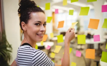 Método SMART ajuda a definir metas profissionais em 2026 Mulher sorrindo em um ambiente de escritório, colando notas adesivas coloridas em uma parede de vidro, ilustrando o processo de organização, visualização e planejamento de metas profissionais descrito na matéria.