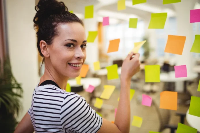Método SMART ajuda a definir metas profissionais em 2026 Mulher sorrindo em um ambiente de escritório, colando notas adesivas coloridas em uma parede de vidro, ilustrando o processo de organização, visualização e planejamento de metas profissionais descrito na matéria.