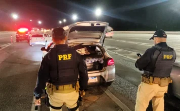 Mortes em rodovias de SC sobem no Carnaval 2026 Policiais rodoviários federais realizam a abordagem e fiscalização de um veículo no acostamento de uma rodovia durante a noite, em Santa Catarina. A imagem ilustra as ações de controle de tráfego da PRF durante o feriado de Carnaval de 2026, mencionado na matéria.