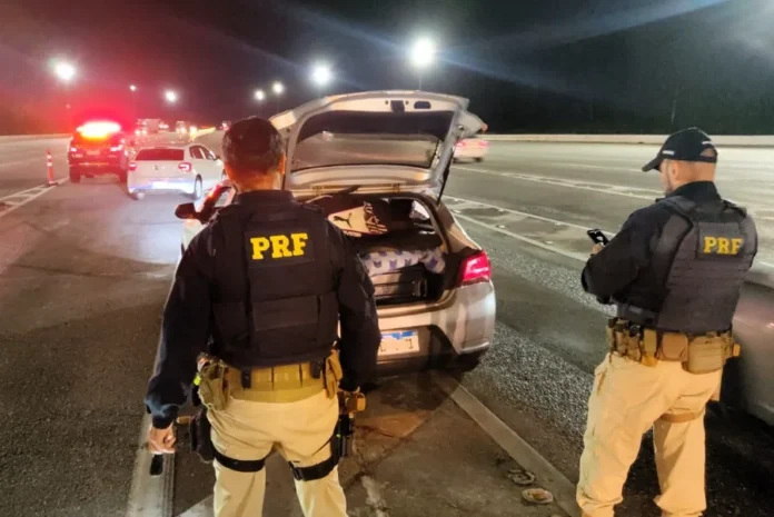 Mortes em rodovias de SC sobem no Carnaval 2026 Policiais rodoviários federais realizam a abordagem e fiscalização de um veículo no acostamento de uma rodovia durante a noite, em Santa Catarina. A imagem ilustra as ações de controle de tráfego da PRF durante o feriado de Carnaval de 2026, mencionado na matéria.