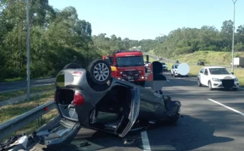 Mulher é resgatada após capotamento em Criciúma "Carro capotado de cabeça para baixo no acostamento da Rodovia Aristides Bolan, em Criciúma, com caminhão do Corpo de Bombeiros ao fundo, relacionado ao acidente descrito na matéria.