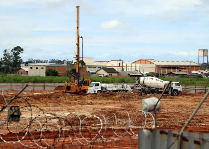 Nova unidade prisional avança em Araranguá Canteiro de obras do novo Presídio Regional de Araranguá em estágio inicial de fundação. A imagem apresenta um terreno de terra batida onde operam máquinas pesadas, incluindo uma perfuratriz amarela de grande porte e dois caminhões-betoneira brancos. Ao fundo, observam-se galpões industriais e residências sob um céu nublado. O primeiro plano é detalhado por uma cerca de segurança com arame do tipo concertina, que contextualiza a natureza da infraestrutura penal em construção.