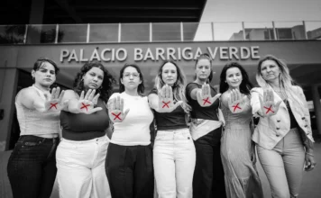 Observatório alerta para alta na violência de gênero em SC Fotografia em preto e branco, com destaque para a cor vermelha, mostrando um grupo de sete mulheres de pé, lado a lado, em frente à fachada do Palácio Barriga Verde, sede da Assembleia Legislativa de Santa Catarina. Com expressões sérias e determinadas, todas estendem uma das mãos para a frente com a palma aberta, exibindo um "X" desenhado em vermelho. O gesto é um símbolo da campanha de combate à violência contra a mulher, tema central da reportagem que aborda os altos índices desse crime no estado.