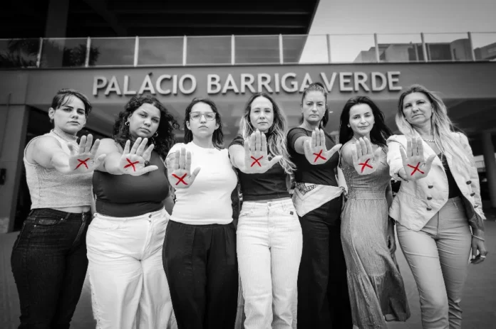 Observatório alerta para alta na violência de gênero em SC Fotografia em preto e branco, com destaque para a cor vermelha, mostrando um grupo de sete mulheres de pé, lado a lado, em frente à fachada do Palácio Barriga Verde, sede da Assembleia Legislativa de Santa Catarina. Com expressões sérias e determinadas, todas estendem uma das mãos para a frente com a palma aberta, exibindo um "X" desenhado em vermelho. O gesto é um símbolo da campanha de combate à violência contra a mulher, tema central da reportagem que aborda os altos índices desse crime no estado.