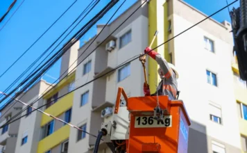 Operação Limpa Fios chega ao Comerciário em Criciúma Técnico de manutenção, utilizando capacete, luvas e colete reflexivo, trabalha dentro de um cesto elevatório laranja para organizar fiação aérea. Ele manipula cabos em um poste de energia em frente a edifícios residenciais, em uma ação de limpeza e remoção de fios inativos durante o dia.