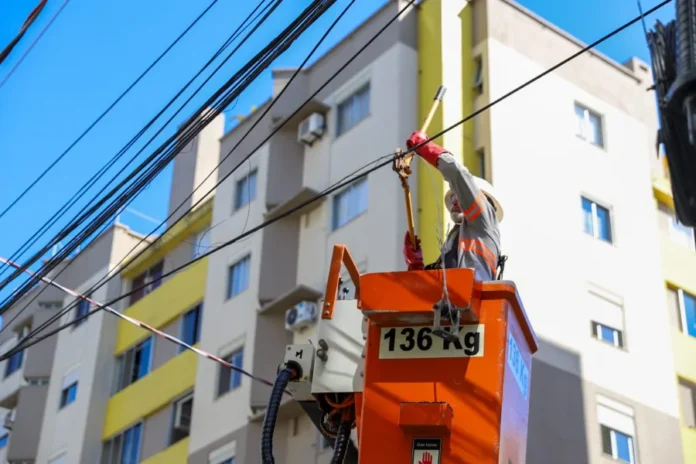 Operação Limpa Fios chega ao Comerciário em Criciúma Técnico de manutenção, utilizando capacete, luvas e colete reflexivo, trabalha dentro de um cesto elevatório laranja para organizar fiação aérea. Ele manipula cabos em um poste de energia em frente a edifícios residenciais, em uma ação de limpeza e remoção de fios inativos durante o dia.