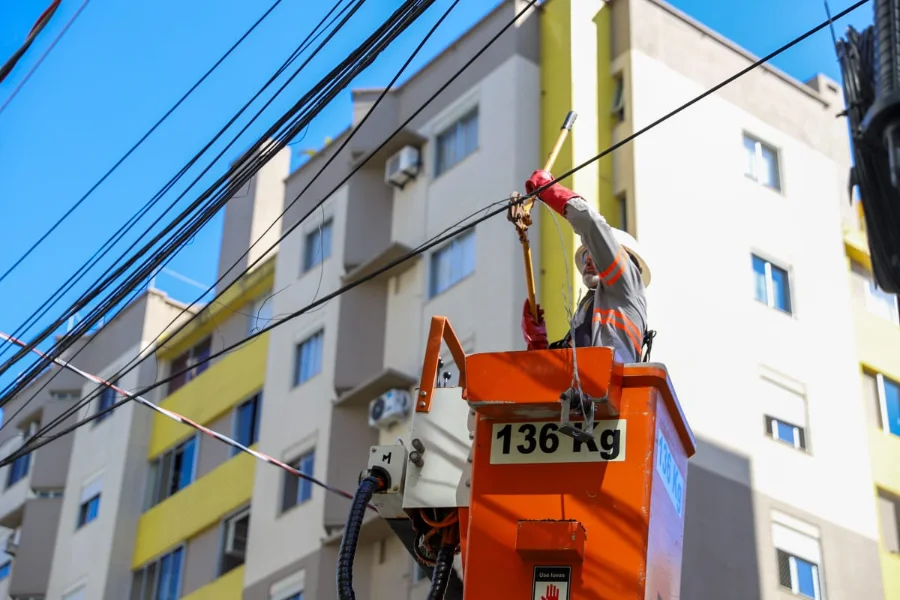 Operação Limpa Fios chega ao Comerciário em Criciúma Técnico de manutenção, utilizando capacete, luvas e colete reflexivo, trabalha dentro de um cesto elevatório laranja para organizar fiação aérea. Ele manipula cabos em um poste de energia em frente a edifícios residenciais, em uma ação de limpeza e remoção de fios inativos durante o dia.