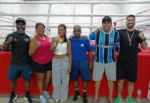 Ouro na Romênia: Boxeadora criciumense é recebida com festa Uma foto de grupo de alta qualidade com seis pessoas na academia de boxe Company em Criciúma. Elas estão em frente a um ringue de boxe com cordas e lona vermelha. A jovem central, Carolina Vargas Pires, segura sua medalha de ouro da Romênia. Atrás deles, um banner de comemoração diz: 'BEM-VINDA, CAROLINA VARGAS PIRES! NOSSA OURO DA ROMÊNIA RUMO AO MUNDIAL!'. O grupo inclui seus pais, técnico e amigos. Há balões e equipamentos de treino ao fundo. Os créditos da foto estão visíveis no canto inferior.