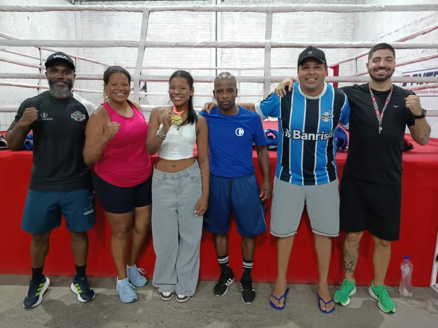 Uma foto de grupo de alta qualidade com seis pessoas na academia de boxe Company em Criciúma. Elas estão em frente a um ringue de boxe com cordas e lona vermelha. A jovem central, Carolina Vargas Pires, segura sua medalha de ouro da Romênia. Atrás deles, um banner de comemoração diz: 'BEM-VINDA, CAROLINA VARGAS PIRES! NOSSA OURO DA ROMÊNIA RUMO AO MUNDIAL!'. O grupo inclui seus pais, técnico e amigos. Há balões e equipamentos de treino ao fundo. Os créditos da foto estão visíveis no canto inferior.