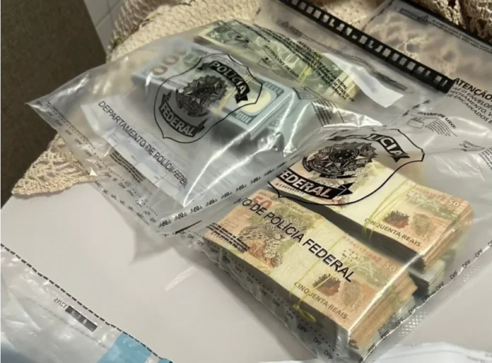 PF desarticula esquema de contrabando de bebidas no Sul Dois envelopes plásticos transparentes, lacrados com o selo oficial da Polícia Federal, contendo grandes maços de dinheiro em espécie. No envelope à esquerda, é possível ver notas de 100 dólares americanos; no da direita, pilhas de notas de 50 reais brasileiros. O material faz parte das apreensões da Operação Carga Dividida, que investiga contrabando e lavagem de dinheiro.