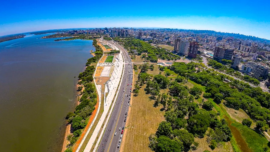 Vista aérea panorâmica da cidade de Porto Alegre, capital do Rio Grande do Sul, mostrando a orla urbanizada do Lago Guaíba, avenidas movimentadas com veículos e a densidade de prédios ao fundo. A imagem ilustra o principal centro urbano do estado, que lidera a expansão econômica nacional em 2026, impulsionada pela agropecuária e com reflexos nos setores de indústria e serviços.