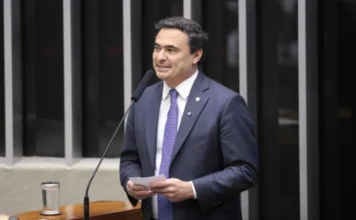 PL aumenta penas para roubo e furto de armas de fogo Fotografia em plano médio do deputado Mauricio Neves (PP-SP). Ele é um homem de pele clara, cabelos escuros e veste um terno azul-marinho com gravata estampada em tons de azul e roxo. O parlamentar está de pé atrás de um púlpito de madeira com um microfone à sua frente, segurando um pequeno papel branco e olhando para o lado como se estivesse discursando. Ao fundo, vê-se a estrutura vertical metálica característica do plenário da Câmara dos Deputados.