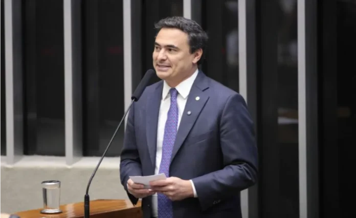 PL aumenta penas para roubo e furto de armas de fogo Fotografia em plano médio do deputado Mauricio Neves (PP-SP). Ele é um homem de pele clara, cabelos escuros e veste um terno azul-marinho com gravata estampada em tons de azul e roxo. O parlamentar está de pé atrás de um púlpito de madeira com um microfone à sua frente, segurando um pequeno papel branco e olhando para o lado como se estivesse discursando. Ao fundo, vê-se a estrutura vertical metálica característica do plenário da Câmara dos Deputados.
