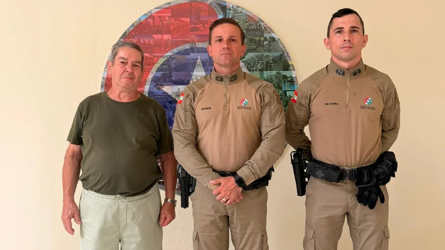 Fotografia mostra um senhor idoso de camiseta verde e calça clara em pé à esquerda, ao lado de dois policiais militares fardados com o uniforme cáqui da Polícia Militar de Santa Catarina. Eles estão posados para a foto dentro de uma sala, com um painel circular colorido ao fundo, registrando o encontro de agradecimento no batalhão.