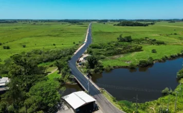 Passo de Torres libera asfalto entre BR-101 e Bellatorres Vista aérea de um longo trecho da nova estrada pavimentada em Passo de Torres. O asfalto escuro e recém-aplicado contrasta com a paisagem rural de campos verdes ao redor. Em primeiro plano, a via atravessa uma pequena ponte sobre um corpo d'água, estendendo-se em linha reta em direção ao horizonte sob um céu azul claro.