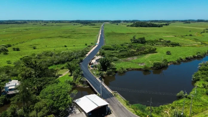 Passo de Torres libera asfalto entre BR-101 e Bellatorres Vista aérea de um longo trecho da nova estrada pavimentada em Passo de Torres. O asfalto escuro e recém-aplicado contrasta com a paisagem rural de campos verdes ao redor. Em primeiro plano, a via atravessa uma pequena ponte sobre um corpo d'água, estendendo-se em linha reta em direção ao horizonte sob um céu azul claro.