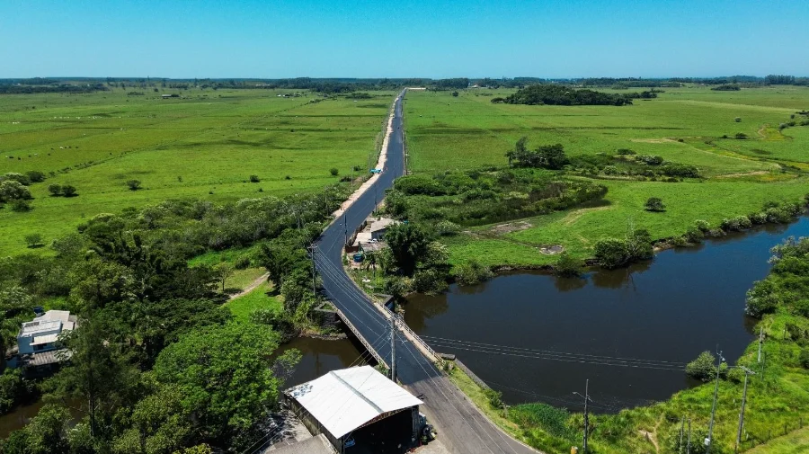 Vista aérea de um longo trecho da nova estrada pavimentada em Passo de Torres. O asfalto escuro e recém-aplicado contrasta com a paisagem rural de campos verdes ao redor. Em primeiro plano, a via atravessa uma pequena ponte sobre um corpo d'água, estendendo-se em linha reta em direção ao horizonte sob um céu azul claro.