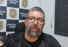 Polícia apreende carro usado em execução em Araranguá Fotografia em plano médio do delegado da Polícia Civil, Lucas Fernandes da Rosa. Ele é um homem de pele clara, tem cabelos curtos escuros, barba cheia e grisalha, e usa óculos de grau com armação preta. Veste uma camiseta preta com a bandeira de Santa Catarina na manga. Ao fundo, vê-se um painel quadriculado com o brasão e a inscrição 'Polícia Civil SC'.