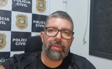 Polícia apreende carro usado em execução em Araranguá Fotografia em plano médio do delegado da Polícia Civil, Lucas Fernandes da Rosa. Ele é um homem de pele clara, tem cabelos curtos escuros, barba cheia e grisalha, e usa óculos de grau com armação preta. Veste uma camiseta preta com a bandeira de Santa Catarina na manga. Ao fundo, vê-se um painel quadriculado com o brasão e a inscrição 'Polícia Civil SC'.