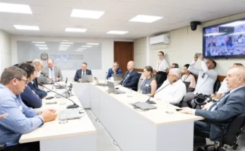 Preço da cebola em SC opera abaixo do custo de produção Foto ampla de uma reunião formal da Comissão de Agricultura e Desenvolvimento Rural na sede da Assembleia Legislativa de Santa Catarina (Alesc). Vários homens vestidos com camisas sociais e paletós estão sentados ao redor de uma grande mesa de conferência em formato de 'U', equipada com microfones e laptops. Ao fundo, uma parede de vidro exibe o brasão e o nome da Assembleia Legislativa. À direita, um monitor grande mostra participantes em videoconferência. O grupo debate medidas para enfrentar os prejuízos na produção de cebola no estado.
