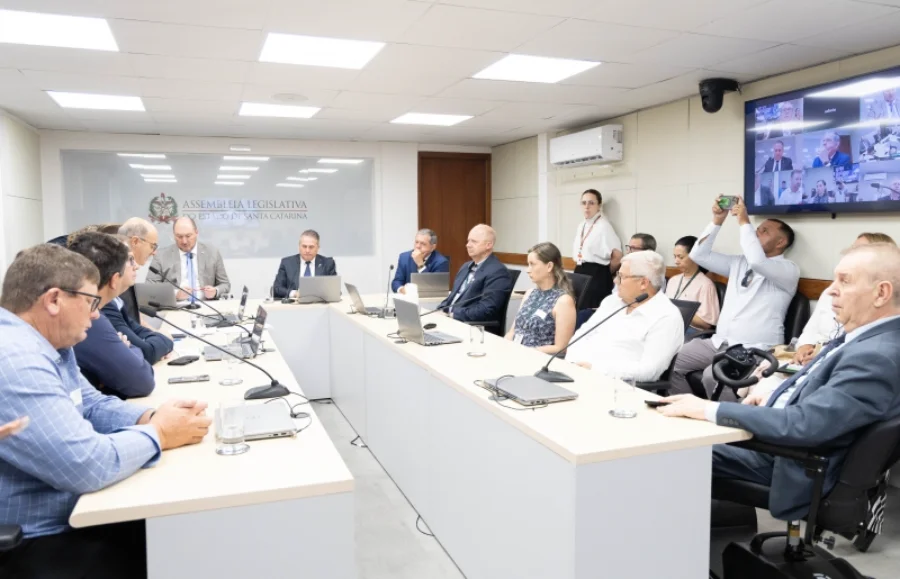 Foto ampla de uma reunião formal da Comissão de Agricultura e Desenvolvimento Rural na sede da Assembleia Legislativa de Santa Catarina (Alesc). Vários homens vestidos com camisas sociais e paletós estão sentados ao redor de uma grande mesa de conferência em formato de 'U', equipada com microfones e laptops. Ao fundo, uma parede de vidro exibe o brasão e o nome da Assembleia Legislativa. À direita, um monitor grande mostra participantes em videoconferência. O grupo debate medidas para enfrentar os prejuízos na produção de cebola no estado.