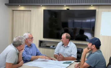 Prefeito de Cocal articula energia para novo distrito industrial Reunião entre o prefeito de Cocal do Sul, Ademir Magagnin, e a diretoria da Cermoful Energia para discutir o projeto de fornecimento de energia elétrica para a 5ª área industrial do município. Os participantes analisam plantas e mapas sobre uma mesa em uma sala de reuniões.