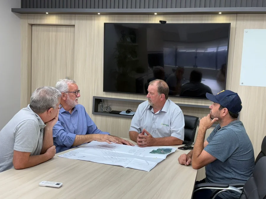 Reunião entre o prefeito de Cocal do Sul, Ademir Magagnin, e a diretoria da Cermoful Energia para discutir o projeto de fornecimento de energia elétrica para a 5ª área industrial do município. Os participantes analisam plantas e mapas sobre uma mesa em uma sala de reuniões.