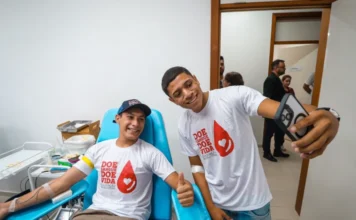 RS alerta para queda nos estoques de sangue no Carnaval Dois jovens sorridentes em um hemocentro. Um deles está sentado em uma cadeira azul de coleta, com o braço estendido realizando a doação de sangue, enquanto o outro está de pé ao lado, segurando um celular para tirar uma selfie de ambos. Eles vestem camisetas brancas com o logotipo de uma gota vermelha e a frase "Doe Sangue, Doe Vida". O ambiente é clínico, com paredes brancas e equipamentos médicos ao fundo.