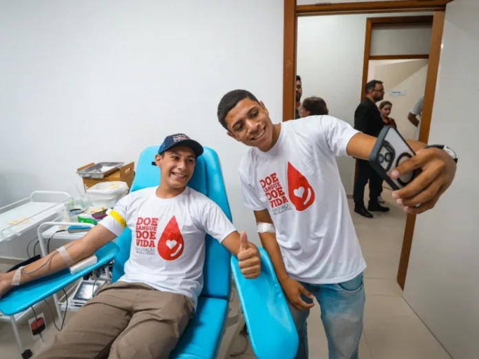 RS alerta para queda nos estoques de sangue no Carnaval Dois jovens sorridentes em um hemocentro. Um deles está sentado em uma cadeira azul de coleta, com o braço estendido realizando a doação de sangue, enquanto o outro está de pé ao lado, segurando um celular para tirar uma selfie de ambos. Eles vestem camisetas brancas com o logotipo de uma gota vermelha e a frase "Doe Sangue, Doe Vida". O ambiente é clínico, com paredes brancas e equipamentos médicos ao fundo.
