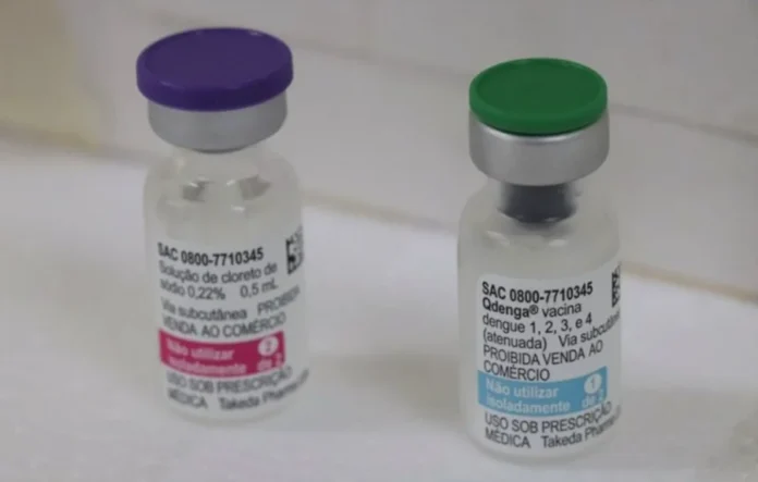 RS amplia vacina contra dengue para todos os municípios Fotografia em close-up de dois frascos da vacina Qdenga sobre uma superfície clara. O frasco à esquerda possui tampa roxa e o da direita possui tampa verde, com o rótulo exibindo o nome do imunizante utilizado na campanha contra a dengue para crianças e adolescentes.