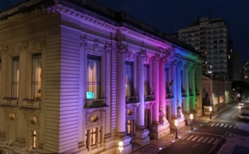 RS inicia semana de conscientização sobre doenças raras Fotografia noturna em ângulo diagonal da fachada lateral e frontal do Palácio Piratini, em Porto Alegre. O prédio histórico de arquitetura clássica está iluminado por projetores de luz nas cores lilás, rosa, azul e verde, cores que simbolizam a campanha do Dia Mundial das Doenças Raras. A iluminação destaca as colunas e detalhes ornamentais da sede do governo, enquanto a rua ao lado aparece com luzes de postes e rastros de veículos ao fundo.